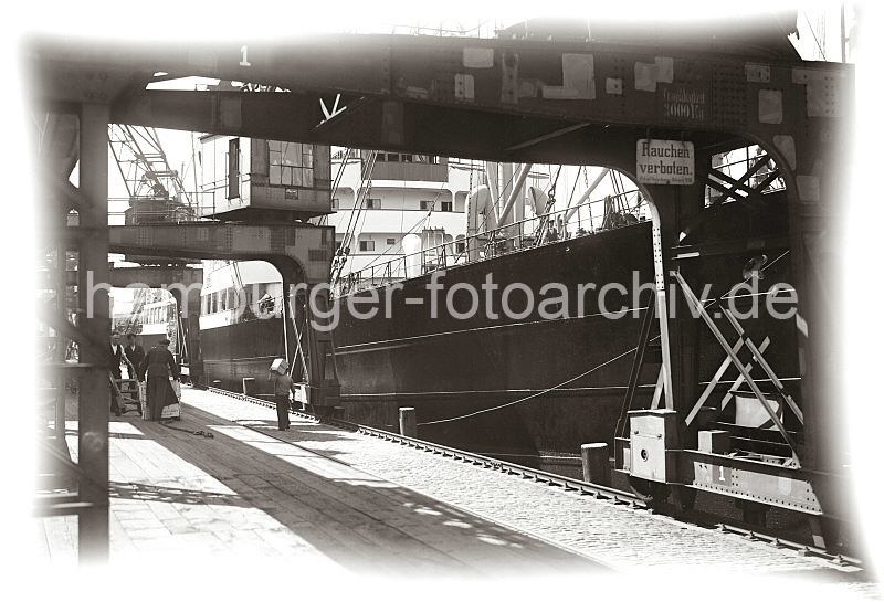 Alte Hafenbilder aus der Hansestadt Hamburg. Arbeit im Hamburger Hafen - Halbportalkrane, Frachter an der Kaimauer; Hafenarbeiter beim Kistentransport.  585_661a Blick durch die mchtigen Halbportale der Hafenkrne am Australiakai des Hamburger Indiahafens; die Krane laufen auf einer Schiene, die direkt an der Kaimauer liegt. Mehrere Arbeiter laden eine Holzkiste fr den Weitertransport auf eine Sackkarre, ein weiterer Hafenarbeiter hat eine kleinere Kiste geschultert und trgt sie ber den Hafenkai.