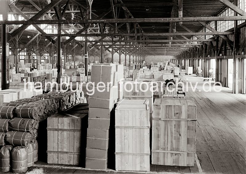 Arbeit im Hafen Hamburgs - Fotos aus dem Archiv der HHLA. Lagerschuppen - gestapelte Gter, Elektrokarren / Sackkarren.  586_662a Die Waren stehen gut und sicher verpackt in einem Lagerschuppen des Hamburger Hafens - im Hintergrund transportieren Hafenarbeiter Kisten mit Elektrokarren. Sackkarren mit den gebogenen Handgriffen sind in den Gngen des Lagers abgestellt.