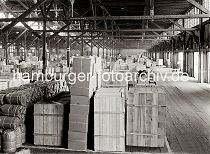586_662a Die Waren stehen gut und sicher verpackt in einem Lagerschuppen des Hamburger Hafens - im Hintergrund transportieren Hafenarbeiter Kisten mit Elektrokarren. Sackkarren mit den gebogenen Handgriffen sind in den Gängen des Lagers abgestellt.
