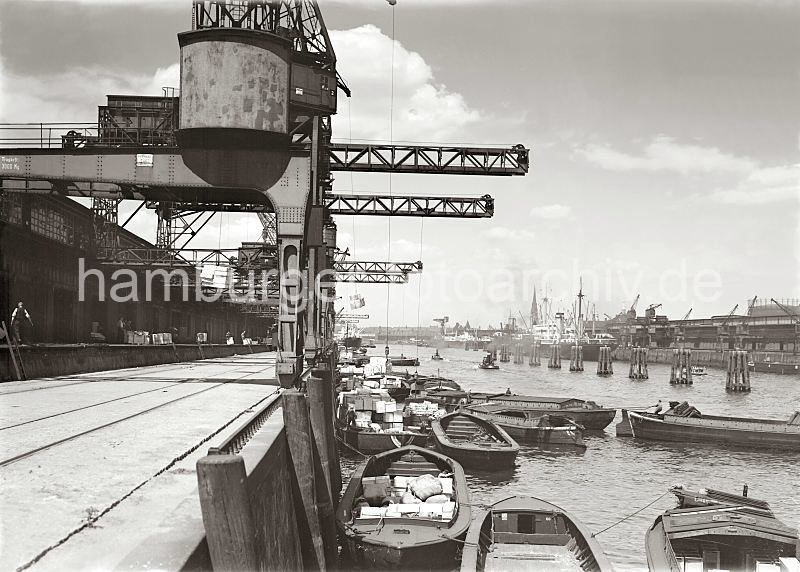 Bilder aus der Geschichte des Hafen Hamburgs - Archiv der HHLA - Beladen  von Schuten am Kamerunkai / Indiahafen - Blick Richtung Norderelbe, Strandhafen. 587_663 Am Kamerunkai des Hamburger Indiahafen liegen Schuten, die ber die Krananlagen mit Kisten und Ballen beladen werden - Lagerarbeiter haben Kisten auf der Laderampe fr die Verladung abgestellt. Zwei Schlepper fahren und dichtem Qualm im Hafenbecken - sie schleppen die antriebslosen Lastkhne durch den Hafen Hamburgs. In der Mitte des Hafenbecken sind Holzdalben in den Hafengrund getrieben, an denen Schiffe festmachen knnen, dahinter hat am Australiakai ein Frachter fest gemacht. Im Hintergrund in der Bildmitte die Schuppen des Strandhafens auf der gegenberliegenden Seite der Norderelbe.