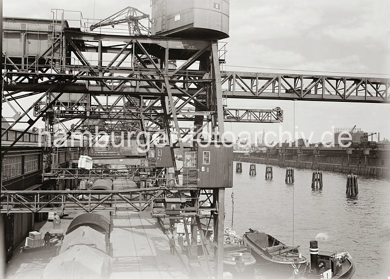 Bilder aus dem historischen Hafen Hamburgs - Krananlagen am Kamerunkai des Indiahafen - Hamburg-Bilder als Geschenke aus Hamburg zu Weihnachten. 588_663a Ein Kran hat gerade eine Last Kisten von der Laderampe des Kamerunkais im Indiahafen aufgenommen und bringt die Ladung zu einer an der Kaimauer liegenden Schuten. Auf der Rampe warten Elektrokarren mit Anhnger, auf denen weitere Kisten stehen. Unter den Portalen der Hafenkrane steht ein Gterzug, das zweite Gleis ist leer.