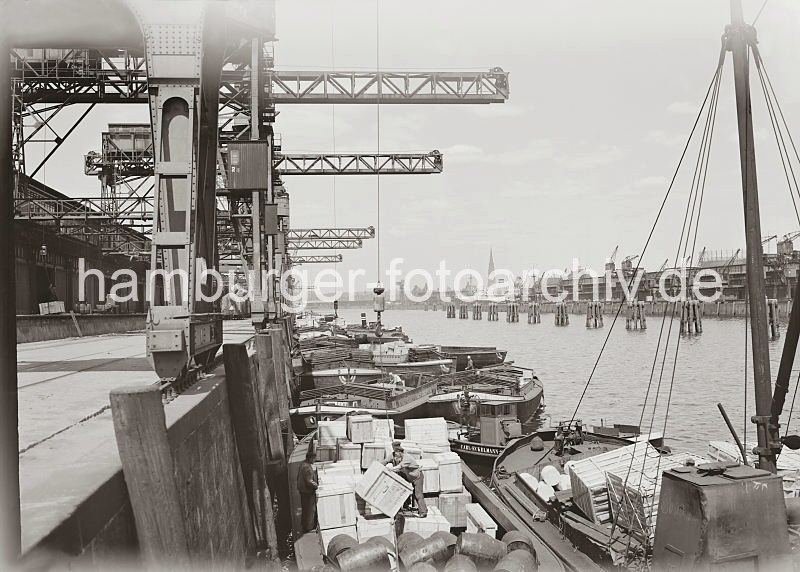Bilder aus dem alten Hafen Hamburgs - Kistenverladung im Indiahafen - Gterumschlag in Schuten. Hamburg-Geschenke: Bilder zu Weihnachten. 589_664 Eine Kiste wird im Indiahafen mit einem Kran an Bord einer Schute gebracht - Hafenarbeiter nehmen die Ladung an und stapeln sie auf eine weitere Kiste. Im Hintergrund ist der Turm der St. Nikolaikirche auf der anderen Seite der Elbe zu erkennen.