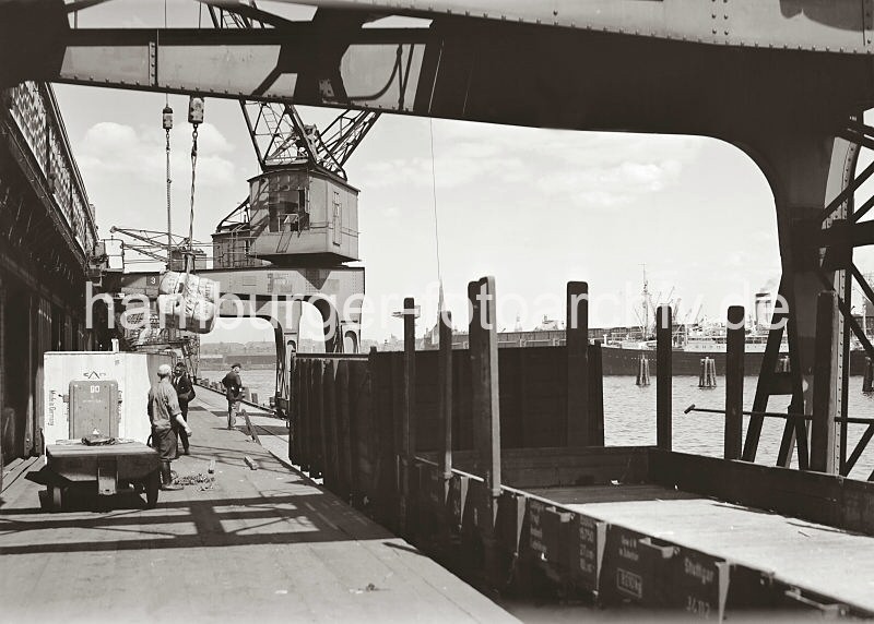 Historische Hafenfotos aus dem Archiv der Hamburger Hafen und Logistik AG HHLA. Elektrokarren auf der Laderampe - Gterwaggons auf der Kaianlage. Hamburgbilder: Fotos als Weihnachtsgeschenke. 591_665a Mit Elektrokarren mit Anhnger bringen die Fracht aus dem Lagerschuppen auf die Laderampe des Kamerunkais im Hamburger Indiahafen. Ein Kran nimmt die Ballen mit dem Ladegeschirr an den Haken, um die Ladung auf die Schuten zu bringen, die an der Kaimauer fest gemacht haben. Im Vordergrund zwei leere Waggons eines Gterzugs an der Rampe.
