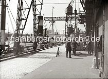 594_650 In Ein Frachtschiff mit hohem Schornstein liegt an einem Kai im Hamburger Hafen, die Ladung des Frachters wird gelscht; gerade wird eine Hieve mit Kisten von einem der Halbportalkrane auf der Laderampe abgesetzt. Im Hintergrund transportieren Hafenarbeiter mit Sackkarren die Gter in den Lagerschuppen.