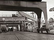 595_B_326 Die hohen Portale der Hafenkrne berspannen die Kaianlage; Pferdefuhrwerke stehen an der Laderampe, die Anhnger sind mit Fruchtkisten beladen. Ein Transportarbeiter deckte die Ladung auf dem Pferdeanhnger mit einer Plane gegen die Klte ab. Die Flagge eines Frachters aus Norwegen / Oslo weht am Heck des Schiffs - ber ein Frderband, das mit Stoff gegen die Witterung geschtzt ist, wird eine Ladung Bananen gelscht und in den Kaischuppen transportiert.