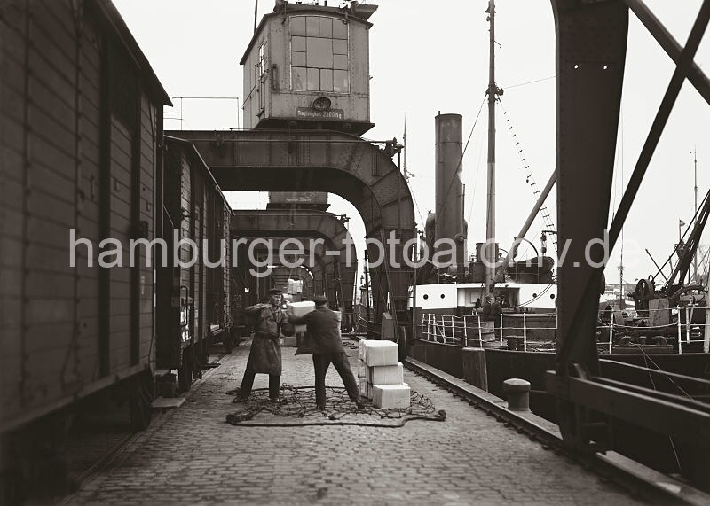 Alte Motive vom Hafen Hamburgs - Fotoarchiv der Hamburger Hafen und Logistik AG / HHLA. Hafenarbeiter beladen einen Gterzug von Hand.  597_554a Zwei Hafenarbeiter stehen auf dem Hafenkai in dem auf dem Boden liegenden Transportnetz und laden die an Land gebrachten Kartons von Hand in den Gterwaggon. Im Hintergrund wird gerade eine Ladung Kartons mit einem der Hafenkrane an Land gebracht.