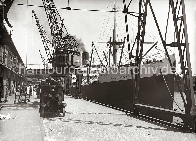 Historische Bilder aus dem Fotoarchiv der Hamburger Hafen und Logistik AG / HHLA; Gterumschlag im Hafen der Hansestadt Hamburg - Lastkraftwagen + Pferdefuhrwerk.  59_650a Gterumschlag am Hafenkai - Kisten mit Frchten werden von den Hafenkranen auf der Laderampe abgesetzt. Mit Handkarren bringen die Arbeiter die Fruchtkisten in den Lagerschuppen. Im Vordergrund steht ein Lastkraftwagen mit geffneter Motorhaube - unter dem Khlergrill befindet sich die Handkurbel. Hinter dem LKW steht ein Pferdefuhrwerk, der Kutscher wartet auf seine Ladung.