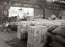 607_673 Blick in den Lagerschuppen 15 am Dalmannkai des Hamburger Grasbrookhafens - Holzkisten und Scke sind in dem Lagerraum gestapelt - Hafenarbeiter schieben leere Sackkarren zur Laderampe. Links weist ein Schild auf Verletzungsgefahr hin, die durch Ballen mit Bandeisen und Draht entstehen kann und dass nicht unter der schwebenden Last gestanden werden soll.