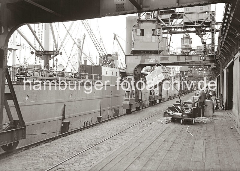 Historische Fotos vom Welthafen Hamburg - Fotoarchiv der HHLA. Lschen eines Frachtschiffs am Dalmannkai, Grasbrookhafen - Halbportalkrane, Ladung Kisten; ca. 1936.  609_675a Eine Ladung Kisten hngt am Kranhaken des Hafenkrans, dessen Halbportal die Kaianlage und Laderampe des Dalmannkais vom Hamburger Grasbrookhafen berspannt. Der Kranfhrer sitzt oben in dem Fhrerhaus, das rundherum verglast ist, damit er einen guten berblick ber den Ladevorgang hat. Auf der Rampe wurden gerade angelandete Kisten abgesetzt.