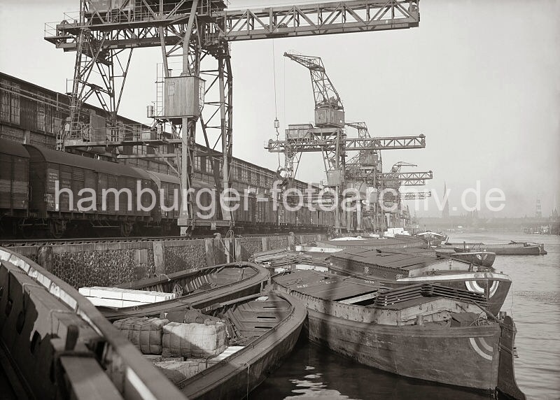 Historische Hafenfotos aus dem Bildarchiv der Hamburger Hafen und Logistik AG, HHLA. Schuten an der Kaimauer des Kamerunkai - Gterzug unter den Hafenkranen. 619_640 Lngs der Kaimauer des Kamerunkais liegen groe Schuten - in ihren Laderumen transportieren die Lastkhne Ballen und Kisten. Bei einigen der Schuten ist der Laderaum mit Holz abgedeckt, um die Ladung gegen die Witterung zu schtzen. Auf der Gleisanlage vor der Laderampe der Kaischuppen steht ein Gterzug - im Hintergrund auf der anderen Seite der Elbe sind die Kirchtrme der Kirchen St. Nikolai, St. Katharinen und St. Petri zu erkennen.