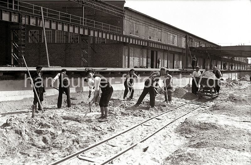 622_12_B_203a Der Hafenkai am Kaischuppen 58 wird gebaut; Bauarbeiter schaufeln Sand - eine mit Sand gefllte Lore wird auf provisorischen Gleisen von drei Mnnern entlang der Laderampe geschoben.  Historische Hafen-Bilder aus dem Fotoarchiv der HHLA. Baustelle am Verteilungsschuppen 57 / 58 - Bauarbeiter mit Schaufeln - Neubau der Kaianlage Togokai; ca. 1934. 