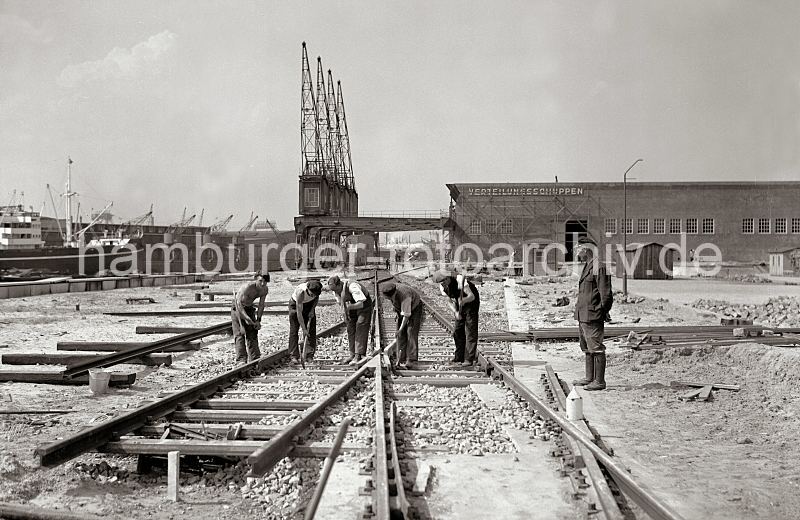 622_13_B_204a Die Gleisanlagen der Hamburger Hafenbahn werden beim Verteilungsschuppen 58 am Togokai des Sdwesthafens verlegt. Bauarbeiter mit Spitzhacke verteilen den Schotter zwischen den Bahnschwellen. Im Hintergrund der Verteilungsschuppen mit dem Schriftzug an der Fassade und vier nebeneinander stehende Hafenkrane, die Ausleger ragen in den Himmel. Historische Hafen-Fotografien aus dem Archiv der HHLA. Baustelle am Verteilungsschuppen 58 - Bauarbeiter verlegen die Gleisanlage der Hafenbahn am Togokai. 