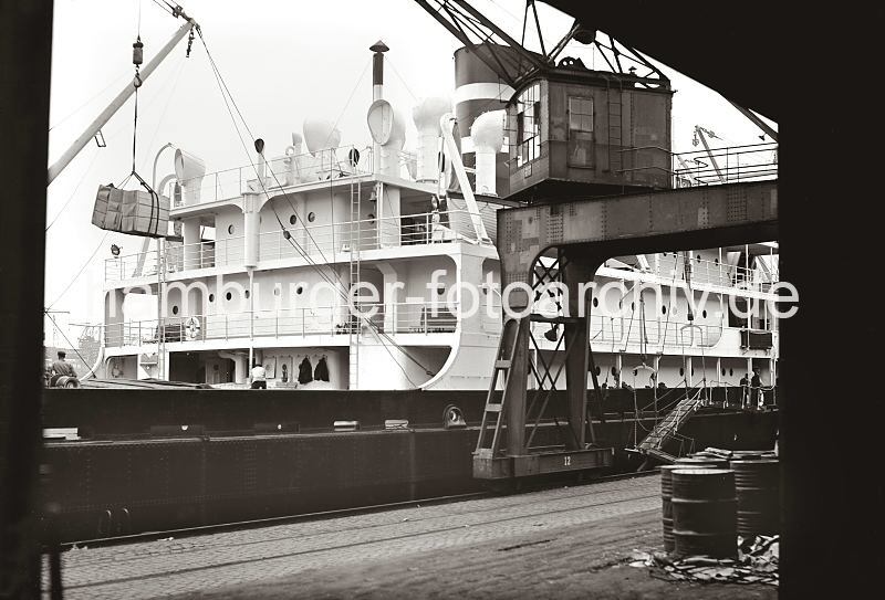 Gterumschlag im Hafen - Fotoarchiv der HHLA - Kisten im Ladegeschirr des Halbportalkrans; Frachtschiff am Hafenkai. 622_18_B_254 Frachter am Kai im Hamburger Hafen; ber seinem Laderaum schwebt eine Ladung Kisten am Haken des Krans. Metallfsser stehen auf der Laderampe des Kaischuppens und warten auf den Abtransport.