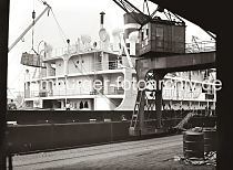 622_18_B_254 Frachter am Kai im Hamburger Hafen; ber seinem Laderaum schwebt eine Ladung Kisten am Haken des Krans. Eine bewegliche Gangway, die den Tidenhub des Wasserstands im Hamburger Hafen ausgleichen kann, fhrt von dem Frachtschiff an Land. Metallfsser stehen auf der Laderampe des Kaischuppens und warten auf den Abtransport.  