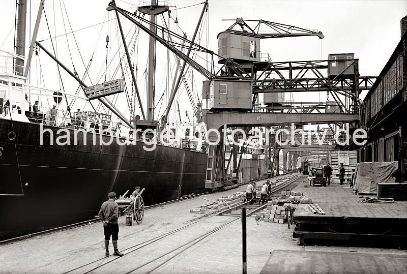 Historische Arbeitsbilder aus dem Fotoarchiv der Hamburger Hafen und Logistik AG - Beladen eines Frachtschiffs - Gleisarbeiten auf dem Hafenkai; ca.1934.  622_19_B_254a Ein Frachtschiff wird im Hamburger Hafen beladen - ein Halbportalkran transportiert eine groe Holzkiste zum Laderaum des Schiffs; weitere Kisten sind auf der Laderampe des Hafenschuppens gestapelt und sollen an Bord gebracht werden. Die Gleisanlage der Hafenbahn, die unter den Halbportalen der Hafenkrane verluft wird repariert - die quadratischen Pflastersteine wurden aus dem Gleisbett entfernt, vier Arbeiter heben eine Eisenschwelle an ihren Platz. Auf dem Kai steht ein Milchhndler(?) mit leeren Flaschen auf der Karre, die Metall-Milchkanne ist auf dem Boden abgestellt.