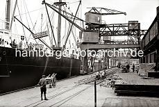 622_19_B_254a Ein Frachtschiff wird im Hamburger Hafen beladen - ein Halbportalkran transportiert eine groe Holzkiste zum Laderaum des Schiffs; weitere Kisten sind auf der Laderampe des Hafenschuppens gestapelt und sollen an Bord gebracht werden. Die Gleisanlage der Hafenbahn, die unter den Halb- portalen der Hafenkrane 