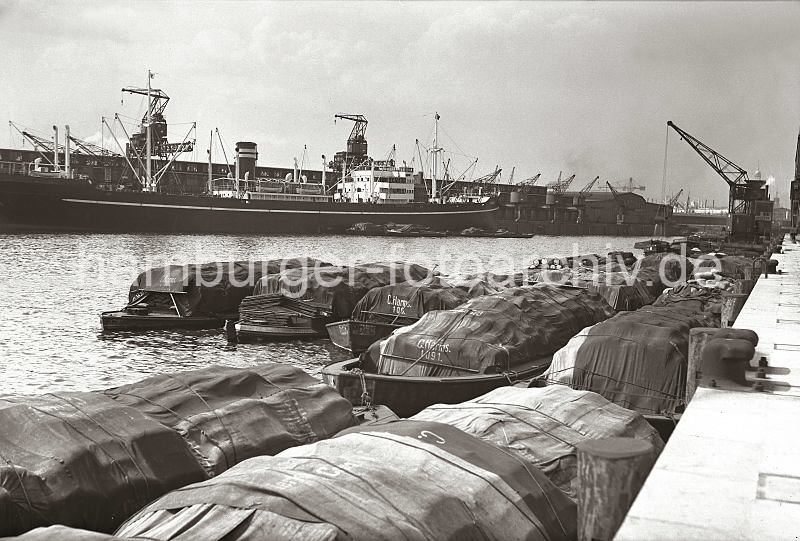 Alte Aufnahmen aus Hamburgs Hafen - Bilder aus dem Archiv der HHLA. Beladene Schuten am Togokai, Frachtschiff am Afrikakai. 622_23_B_208a Bis zu fnf Reihen tief liegen die hoch beladenen Schuten am Togokai des Hamburger Sdwesthafens. Die Ladung, die aus prall gefllten Scken und Tonnen besteht, ist zum grten Teil gegen die Witterung mit Planen geschtzt. Im Hintergrund scheint ein Schwimmbagger das Hafenbecken auszubaggern und den Schlick auf einer Schute abzuladen. Auf der gegenberliegenden Seite des Sdwesthafens liegt der Frachter TUDOR am Afrikakai.