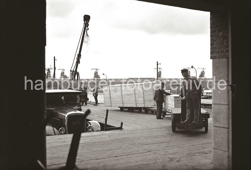Alte Fotos aus dem Hamburger Hafen. Henschel LKW an Laderampe - Transport von Holzkisten mit Elektrokarren + Anhnger.  622_27_B_253 Ein Henschel Lastwagen mit dem Markenzeichen, einem Stern und den Buchstaben HuS / Henschel und Sohn am Khler, steht an der Laderampe des Hafenschuppens 58. Der Transportarbeiter fhrt die Elektrokarre mit den gerade abgeladenen Holzkisten auf den Anhngern in das Innere des Kaischuppens. Im Hintergrund sind die Dcher des Schuppen 59 am Windhukkai des Hamburger Sdwesthafens zu erkennen.