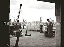 622_27_B_253 Ein Henschel Lastwagen mit dem Markenzeichen, einem Stern und den Buchstaben HuS / Henschel und Sohn am Khler, steht an der Laderampe des Hafenschuppens 58. Der Transportarbeiter fhrt die Elektrokarre mit den gerade abgeladenen Holzkisten auf den Anhngern in das Innere des Kaischuppens. Im Hintergrund sind die Dcher des Schuppen 59 am Windhukkai des Hamburger Sdwesthafens zu erkennen.