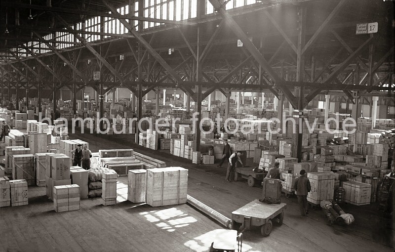 Historische Bilder von der Arbeit im Hafen - Archiv der HHLA. Verteilungsschuppen 57 - Lagerung von Kisten, Elektrokarren; ca. 1936.  626_B_206 Blick in den groen Verteilungsschuppen 57 des Hamburger Indiahafens. Holzkisten sind dort gelagert, die auf ihren Weitertransport warten. Die Anhnger eines Elektrokarrens sind beladen, ein anderer fhrt leer durch den Gang. Die einzelnen Abteilungen des Lagers sind unter dem Dach nummeriert, neben der 17 oben rechts ein Schild mit den Lndernahmen LETTLAND, ESTLAND, RUSSLAND.