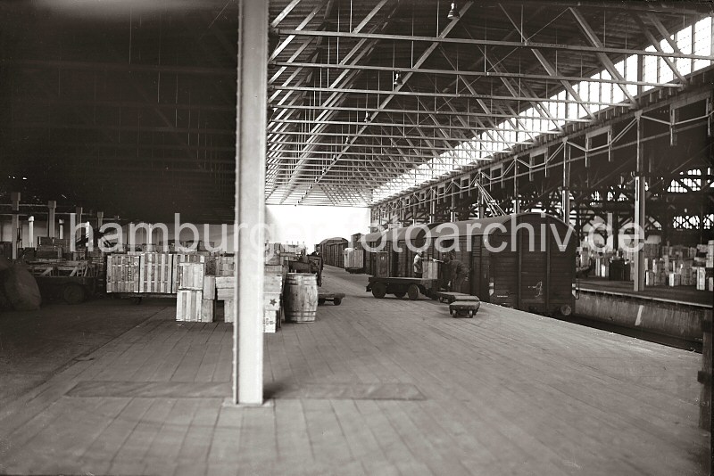Historische Fotografien von der Arbeit im berseehafen Hamburg aus dem Archiv der HHLA. Gterwaggons im Lagerschuppen - Lagergut, Elektrokarren mit Anhnger; ca. 1936. 626_B_206 Geschlossene Gterwaggons stehen im der Mitte des Lagerschuppens, in dem die Ware zumeist in Holzkisten  zum Schutz beim Transport sicher verpackt ist. Ein leerer Elektrokarren  und Lagerarbeiter stehen vor einem der gedeckten Gterwagen -  ber einem der Puffer ist das Schild AUSTAUSCHBAU angebracht.