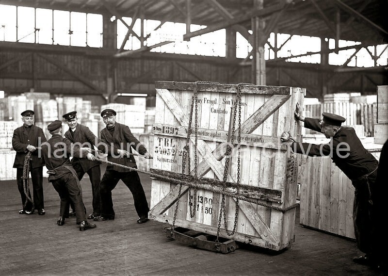 Historische  Arbeitsbilder aus dem Hafen Hamburgs - Bildarchiv der HHLA: Schwertransport einer Holzkiste per Schwerlastrollern - Hafenarbeiter im Hamburger Hafen; ca. 1932. 631_67 Fnf Mann arbeiten daran eine groe Holzkiste mit Glasscheiben durch das Lager im Hamburger Hafen zu transportieren. Die Kiste steht auf Schwerlastrollen und ist mit einer Eisenkette an dem Gestell fixiert. Vier Mnner ziehen die Ladung mit einem Seil, ein weiterer Lagerarbeit schiebt und hlt die Balance der Kiste.
