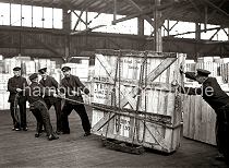 631_67 Fnf Mann arbeiten daran eine groe Holzkiste mit Glasscheiben durch das Lager im Hamburger Hafen zu transportieren. Die Kiste steht auf Schwerlastrollen und ist mit einer Eisenkette an dem Gestell fixiert. Vier Mnner ziehen die Ladung mit einem Seil, ein weiterer Lagerarbeit schiebt und hlt die Balance der Kiste.