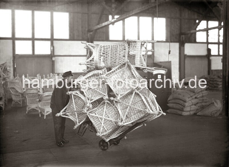 Alte Fotografien von der Arbeit im Hamburger Hafen aus dem - Bildarchiv der HHLA. Transport einer Ladung Korbsthle mit einer Sackkarre - Geschichte der Arbeit im Hafen Hamburgs; ca. 1932.  632_424 Ein Lagerarbeiter hat eine Vielzahl von Korbsthlen auf seine Karre gestapelt und fhrt diese durch den Lagerschuppen im Hamburger Hafen. Im Hintergrund rechts stehen weitere unverpackte Sessel; rechts sind Scke bei einer Waage gestapelt.