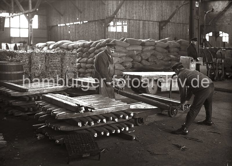 Beladen des Anhngers eines Elektroschleppers mit Kupperbarren; ca. 1932 634_53 Von einem Stapel Kupferbarren haben zwei Arbeiter mit Handhaken einen der Metallbarren aufgenommen und legen diesen auf den Anhnger eines Elektrokarrens. Im Hintergrund steht der Fahrer des Elektroschleppers auf seinem Fhrerstand, einer der Anhnger ist schon beladen.