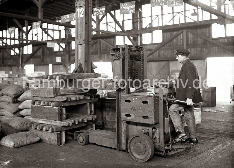 Mit einer Hubkarre werden Kupferbarren in einem Lagerschuppen des Hamburger Hafens transportiert; ca. 1932. 635_71 Mit einer Hubkarre stapelt der Fahrer Kupferbarren im Lager des Hamburger Hafens; auf der Gabel des Hubwagens der Marke YALE liegen mehrer Barren, die auf dicken Bohlen abgelegt werden. Der Fahrer der Elektromaschine bewegt diese mit einem Handhebel und einem Fupedal.