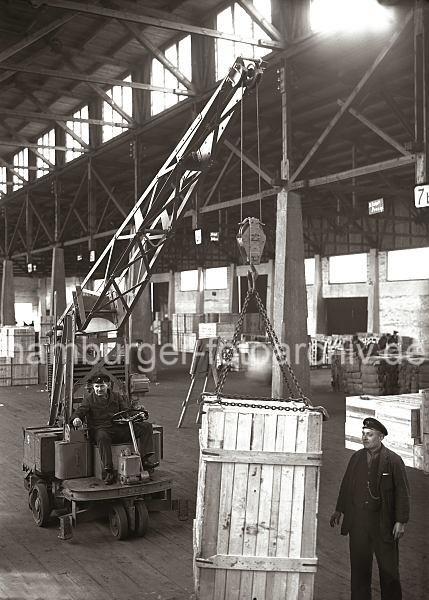 Transport einer Holzkiste mit einem Elektrokran; ca. 1932. Alte Fotografien von der Arbeit im Hamburger Hafen aus dem - Bildarchiv der HHLA. 638_58 Eine groe Holzkiste wird in einer der vielen Lagerhallen des Hamburger Hafens mit einem Laufkran transportiert. Vier Metallkrallen, die sich an einer Eisenkette des Kranhakens befinden, tragen die Kiste, die grob aus Holzbrettern zum Schutz des Transportguts zusammengezimmert ist. Der Fahrer des Elektrokrans hat eine Hand am Lenkrad, whrend die andere auf den Hebeln liegt, mit denen die Kiste am Haken bewegt wird. Ein weiterer Arbeiter dirigiert den Transport zu seinem Ziel.