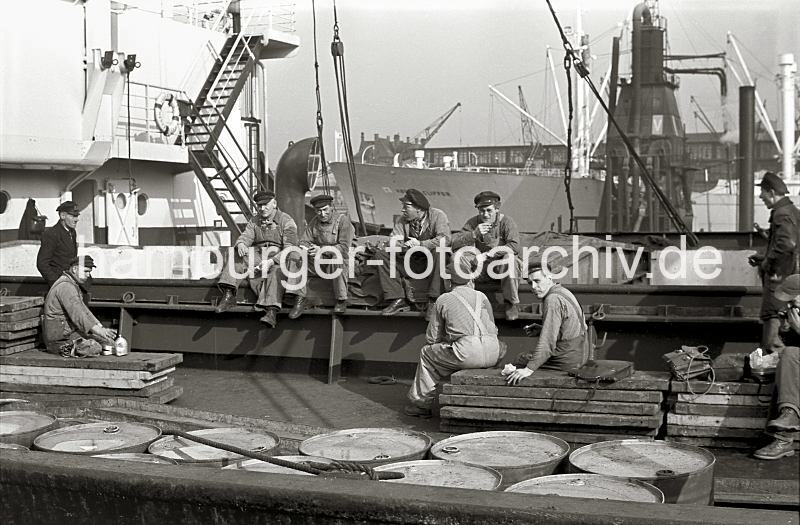 Historische Motive von der Arbeit im Hamburger Hafen aus dem Archiv der HHLA. Hafenarbeiter an Bord eines Frachters machen Pause - Getreideheber; ca. 1954. 641_7 Die Schauerleute, deren Arbeit  im Stauen bzw. dem Be- und Entladen der Frachschiffe besteht machen Pause. Sie essen Brote, die sie in Aktentaschen mitgebracht haben. Einer der Hafenarbeiter greift gerade zu seiner Thermoskanne, der sogen. Kaffeeteng, die er neben seinem Zampel liegen hat. Der Zampel ist ein kleiner Jutesack, in dem der Schauermann sein Essen mit gebracht hat. Der Begriff Schauermann stammt vom niederlndischen Wort sjouwen = schleppen; die Arbeiter im Hafen Hamburgs waren hufig auch Tagelhner, die zur Erledigung einer konkreten Arbeit eingestellt wurden.  Mnner tragen einen "Elbsegler", die typische Hamburger Schirmmtze mit Kordel. Im Hintergrund ist ein Getreideheber zu erkennen.