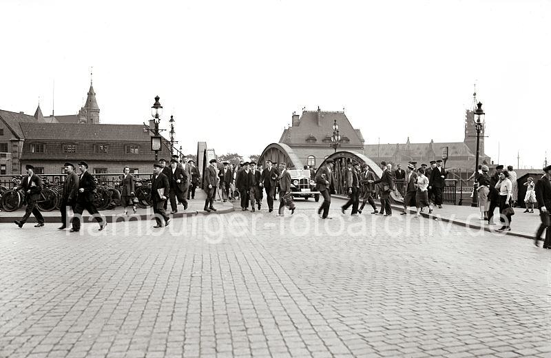 Historische Hamburgfotografien aus dem Archiv der HHLA. Feierabend in der Hamburger Speicherstadt - Arbeiter und Angestellte bei der Niederbaumbrcke. 643_B_280a Es scheint Feierabend zu sein bei den Angestellten und Arbeitern in der Speicherstadt. Mnner und Frauen gehen ber die Niederbaumbrcke, die ber den Zollkanal fhrt. Ein Schild an einer der Laternen weist auf den Zollgrenz-Bezirk hin. Einige Frauen mit Kindern warten auf ihre Mnner - im Hintergrund hinter den eisernen Brckenbogen ist das Dach der Polizeiwache zu erkennen, rechts der Kaispeicher A und lks. einer der Giebeltrme der Speicherstadt.