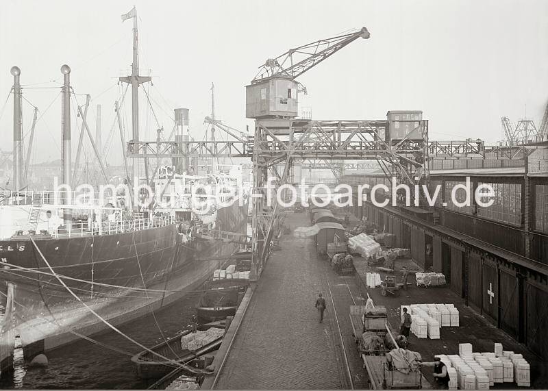Fotos zur Geschichte des Hamburger Hafens - historische Bilder aus dem Archiv der HHLA. Abgebumter Frachter am Kai - Pferdefuhrwerke und Gterwaggons an der Laderampe; ca. 1934. 661_584 Mit Holzstmmen wird der Frachter auf Distanz zur Kaimauer gehalten (abgebumt), so dass die Schuten im Zwischenraum liegen und dort beladen werden knnen. Ballen mit Jute und Baumwolle werden mit den Halbportalkrnen an Land gebracht und auf der Laderampe abgesetzt. Dort stehen mehrere Pferdefuhrwerke, die beladen werden sollen - im Hintergrund geschlossene Gterwaggons an der Rampe.