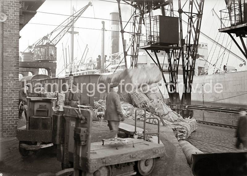 Kaiarbeiter im Hamburger Hafen bei ihrer Arbeit - Elektrokarren mit Anhnger; ca. 1934. 664_586a Hafenkrne lschen die Ladung eines Frachtschiffs - gebndelte Hute liegen auf dem Kaipflaster, eine Hieve mit Wollballen wird gerade auf der Laderampe abgesetzt. Im Vordergrund stehen Elektrokarren mit Anhnger, die auf die Beladung warten. 