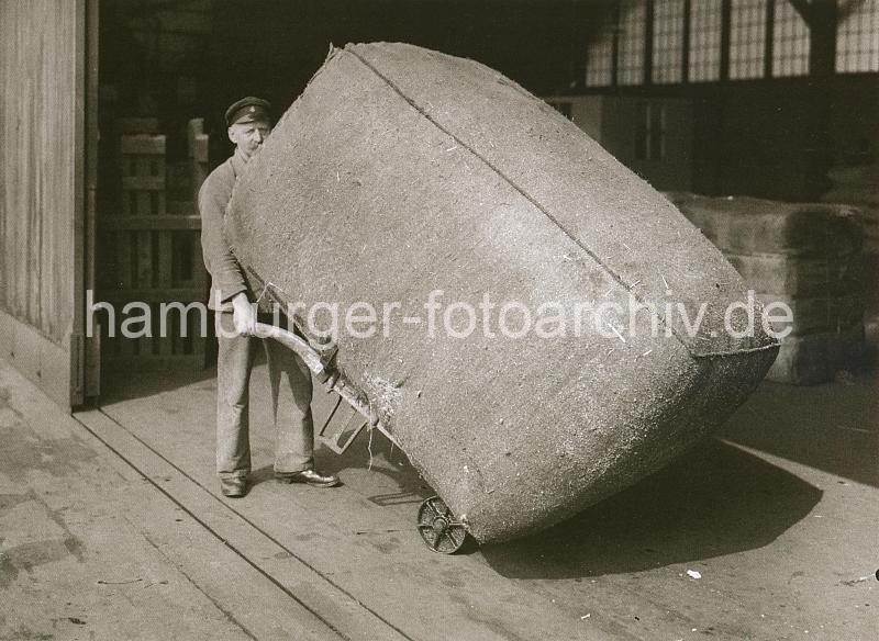 Hafengeschichte in Fotos aus dem Archiv  der Hamburger Hafen und Logistik AG (HHLA) - Handarbeit im Hafen - Transport eines Ballen mit der Sackkarre; ca. 1934.  674_309_421 Ein groer Ballen wird von einem Hafenarbeiter von Hand mit Hilfe einer Sackkarre in den Kaischuppen transportiert.