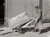 675_426 Der Kaiarbeiter transportiert einen Ballen, der mit Bandeisen zusammengehalten wird auf seiner Sackkarre. Der Hafenarbeiter trägt seine Dienstmütze und eine Schürze zum Schutz seiner Kleidung.