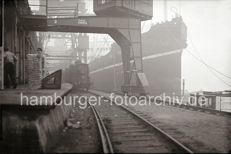 Historische Aufnahmen der HHLA vom Gterumschlag im Hamburger Freihafen. Unter den Portalen der Halbportalkrne steht ein Gterzug an der Rampe - Kaiarbeiter / Frachtschiff; ca. 1934. 687_B_327 Hafenarbeiter transportieren abgeladene Wollballen von der Rampe in den Kaischuppen zur Lagerung. Unter den Portalkrnen steht ein Gterzug an der Laderampe - der hohe Bug eines Frachters ist im Nebel des Hamburger Hafens am Kai zu erkennen.