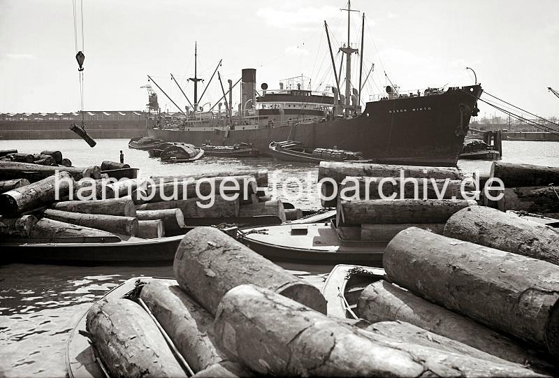 Historische Fotografien zur Hafengeschichte Hamburgs aus dem Archiv der HHLA. Tropische Baumstmme werden auf Schuten geladen; ca. 1938.  692_395a Die Stmme tropischer Bum werden auf Schuten geladen - einer der Baumstmme hngt in einer Baumzange am Kranhacken und wird auf den Lastkahn herunter gelassen. Im Hintergrund liegt das Frachtschiff BARON MINTO an den Holzdalben; die Ladung wird mit schiffseigenen Ladebumen auf auf Schuten gelscht .