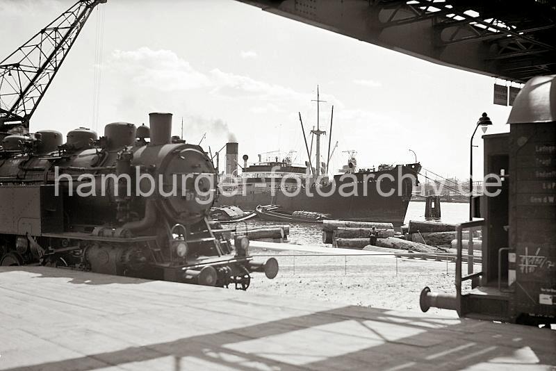 Fotografien zur Geschichte des Hamburger Hafens aus dem Archiv der Frachter und Schuten mit Baumstmmen - Lokomotive, Gterwaggon an der Laderampe;  ca. 1938. 695_396 Blick von der Laderampe auf Schuten, die hoch mit Baumstmmen tropischer Hlzer beladen sind - ein Mann mit Hut und Anzug steht auf der Ladung Holz und macht sich Notizen. Im Hintergrund wird die Ladung eines Frachters gelscht - eine lngsseits liegende Schute wird mit Scken beladen.