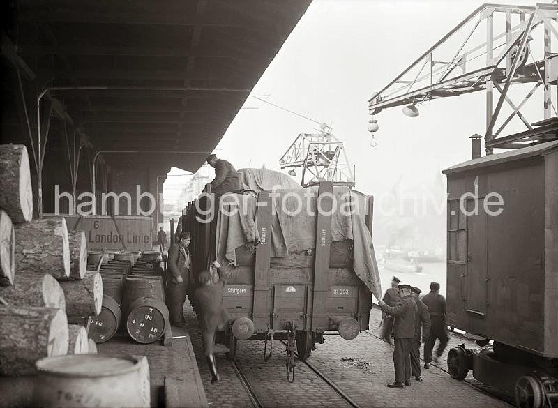Fotoaufnahmen zur Geschichte des Hamburger Hafens aus dem Archiv der HHLA. Kaiarbeiter decken die Ladung eines Gterwaggons miti einer Plane ab;  ca. 1938.  696_427 Vor der Rampe des Kaischuppens steht ein voll beladener offener Gterwaggon - Kaiarbeiter sind dabei die Ladung mit einer Plane abzudecken. Auf der Laderampe sind dicke Baumstmme gestapelt und Fsser abgestellt. Zwei Rollkrne, die auf Schienen laufen stehen in Ruhestellung auf der Kaianlage des Hamburger Hafens.