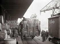 696_427 Vor der Rampe des Kaischuppens steht ein voll beladener offener Gterwaggon - Kaiarbeiter sind dabei die Ladung mit einer Plane abzudecken. Auf der Laderampe sind dicke Baumstmme gestapelt und Fsser abgestellt. Zwei Rollkrne, die auf Schienen laufen stehen in Ruhestellung auf der Kaianlage des Hamburger Hafens.