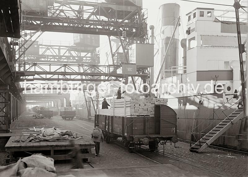 Fotodokumentation zur Arbeit im Hamburger Hafens - Bilder aus dem Archiv. Beladen  eines Frachtschiffs mit Holzbrettern.  699_585a Mit Holzbohlen beladene Gterwaggons sind am Kai auf den Gleisen vor den Frachter geschoben worden - mit dem Kran werden die Bndel Holzbretter von den offenen Waggons gehoben und auf das Schiff transportiert.
