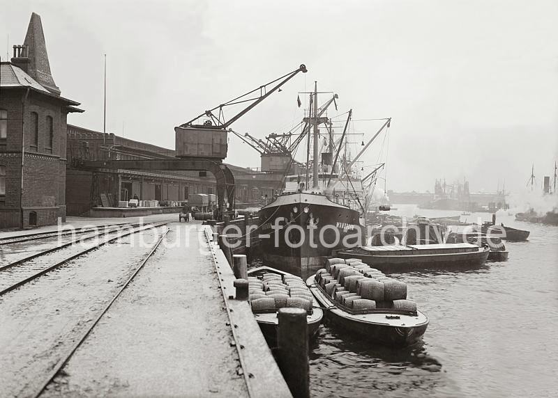 Historische Hafenfotos aus Hamburg - Bilddokumente aus dem Archiv HHLA. Frachtschiff + Schuten am Hafenkai.  702_702a Zwei mit Holzfssern beladene Schuten liegen am Hafenkai - weitere Lastkhne haben auf der Wasserseite des Frachter fest gemacht. Zwei Fsser hngen an dem Ladebaum des Hamburger Frachters WANDRAHM. Tonnen und Holzkisten liegen zum Abtransport auf der Laderampe des Kaischuppens.