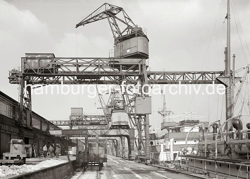 Historische Bilder aus dem Fotoarchiv der Hamburger Hafen und Logistik AG Kaianlage mit Halbportalkrnen im Hafen Hamburgs.  705_728 Unter den Halbportalkrnen stehen offene Gterwaggons auf dem Gleis; auf der Laderampe stehen Kaiarbeiter mit den Hnden in den Taschen im Schnee und warten auf die nchste Ladung. Ihre Sackkarren haben die Hafenarbeiter an der Schuppenwand abgestellt. Lks. im Vordergrund eine Elektrokarre mit leerem Anhnger.