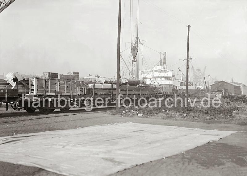 06_545a Auf den Gleisen der Hamburger Hafenbahn stehen zwei hoch mit Kisten beladene offene Gterzge; am Kai sind die Aufbauten eines Frachtschiffs zu erkennen. Im Vordergrund hngt der Haken eines Krans. Historische Fotos aus dem Bildarchiv der Hamburger Hafen und Logistik AG HHLA) Gleisanlage mit beladenen offenen Gterzgen am Hafenkai;  ca. 1934. 