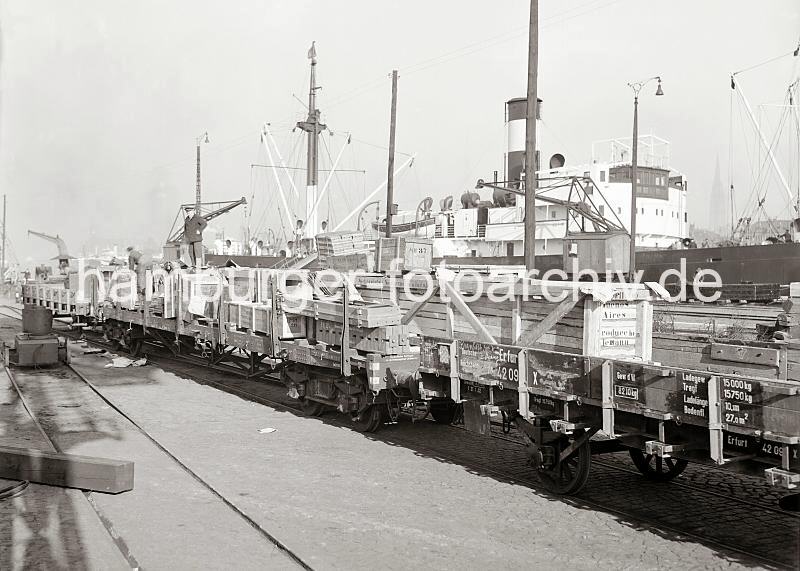707_545b Beladenen Gterwaggons stehen am Holthusenkai; auf den Holzkisten sind die Aufschriften des Bestimmungsortes der Gter BUENOS AIRES sowie die Herkunft PRODUCCION ALEMANA zu erkennen. Zwei Hafenarbeiten stehen auf der Ladung und bereiten die Verladung auf das Schiff vor; lks. steht auf dem Gleis ein Schleppwagen, mit dem ein Gterwaggon ohne die Hilfe einer Lokomotive von einem Arbeiter bewegt werden kann.  Hinter den Schiffsaufbauten ist rechts der Turm der Nikolaikirche zu erkennen. Historische Fotos aus dem Bildarchiv der Hamburger Hafen und Logistik AG (HHLA)Beladene Gterzugwaggons / Frachter am Hafenkai ca. 1934. 