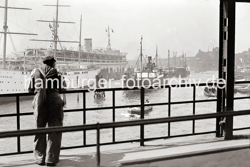 Historische Fotos zur Geschichte des Hamburger Hafens - Blick von der berseebrcke auf den Niederhafen und zu den St. Pauli Landungsbrcken.  711_386 Ein Hafenarbeiter steht am Gelnder der berseebrcke und blickt auf die Schiffe im Hamburger Niederhafen. Einige der Frachter liegen an den Holzdalben, andere Schiffe an der Kaimauer der Rhein Maas Schuppen am Johanisbollwerk. Links das Jugendherbergsschiff HEIN GODENWIND und das KdF Passagierschiff Wilhelm Gustloff. Im Hintergrund die Kuppeln der St. Pauli Landungsbrcken.