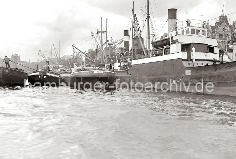 Fotos zur Hamburger Hafengeschichte aus dem Archiv HHLA Ewerfhrer mit Bootshaken - Schuten am Johannisbollwerk; ca. 1934. 716_3_379 Motorschiffe liegen am Kai des Johannisbollwerks - lngsseits haben Lastkhne fest gemacht auf die die Gter verladen werden oder Ladung fr den Weitertransport aufgenommen wird. Ein Ewerfhrer bewegt seine Schute, indem er sie mit dem langen Peekhaken / Bootshaken einem anderen Boot abstt. Dahinter der Pegelturm der Landungsbrcken und das Dach des Fhrhau St. Pauli und der Navigationssschule.