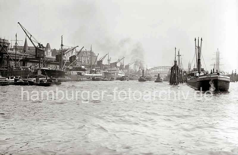 Historisches Hafenpanorama vom Hafen - Rheinschuppen und Hafenkrne an den Vorsetzen - Binnenschiff an den Dalben.  735_387a Hafenanlagen der Rhein / Maas Binnenschiffe an den Vorsetzen. Kstenmotorschiffe liegen am Kai - ihre Ladung wird mit den Krnen an Land gebracht oder auf die wasserseitig liegenden Kleinschiffe wie Schuten oder Barkassen abgegeben. Ein Hafenschlepper zieht einen Elbkahn ber das Wasser - rechts macht ein Binnenschiff an den Dalben fest. Ein Fastmoker (Festmacher) ist auf die Holzstmme gestiegen und vertut das Schiff - im Hintergrund die berseebrcke.